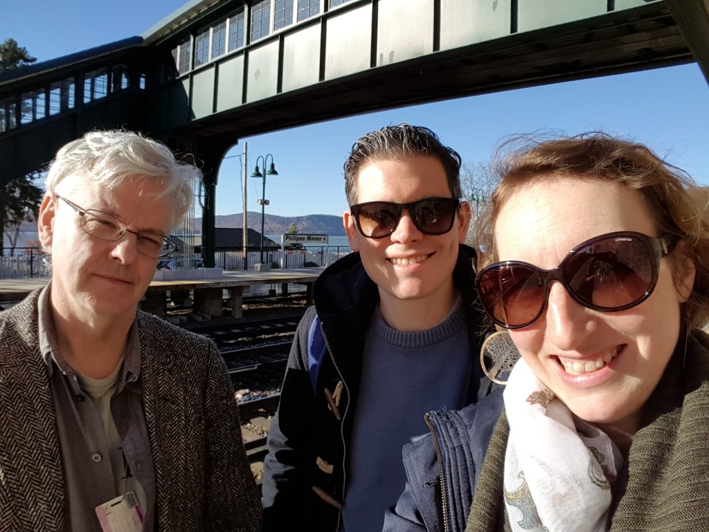 Seamus, Paul and Anna standing on a balcony overlooking rail tracks and the Hudson River. It is a sunny day.